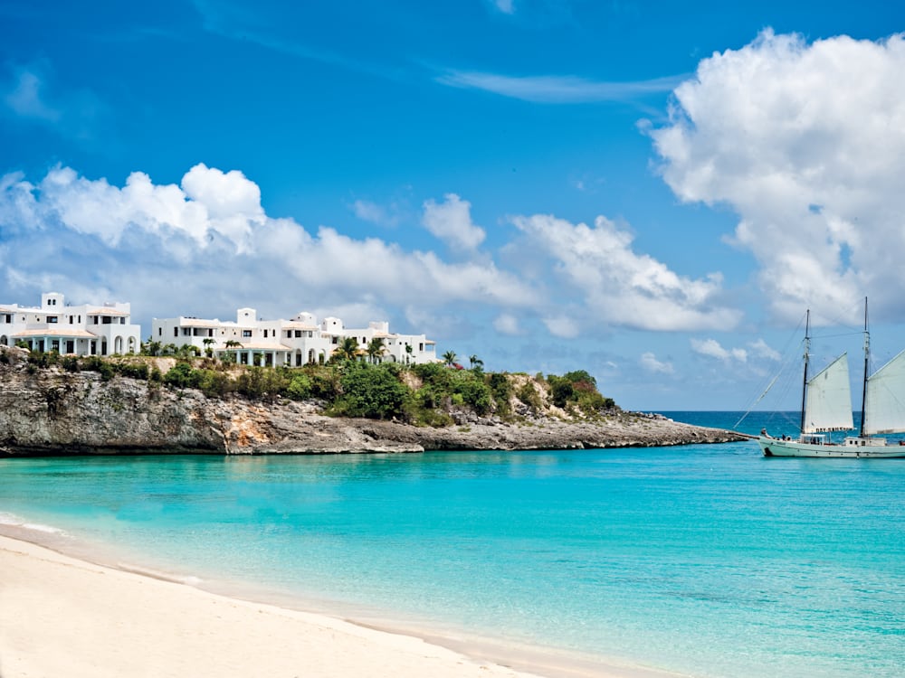 Вид на белый отель на вершинах утесов с белого песчаного пляжа, на лазурно-голубых водах отдыхает парусник.