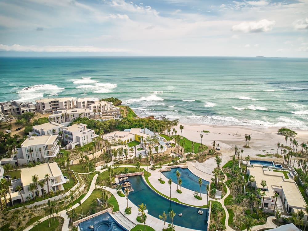 Вид с воздуха на отель и бассейны с видом на волны, катящиеся по белому песчаному пляжу.